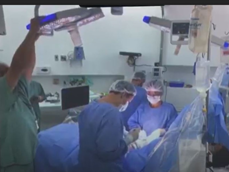 Foto da sala de cirurgia. Dois médicos de roupa azul estão operando a paciente, um de cada lado da mesa. Uma pessoa também de azul aparece ao fundo, de costas, e outra pessoa está ajustando um aparelho no alto, do lado esquerdo